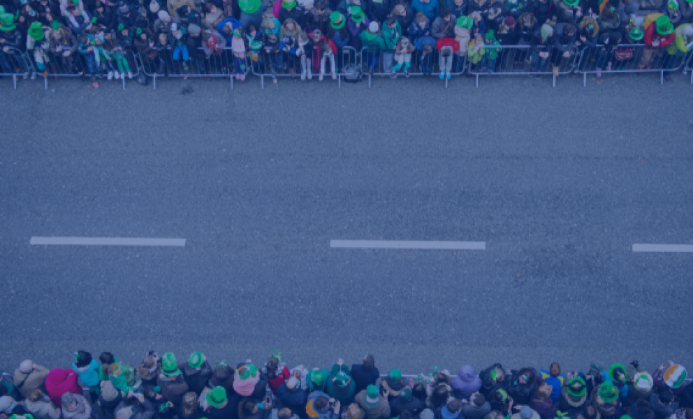 A road prepared for a parade. People gathered either side behind barriers wearing Irish themed paraphernalia 