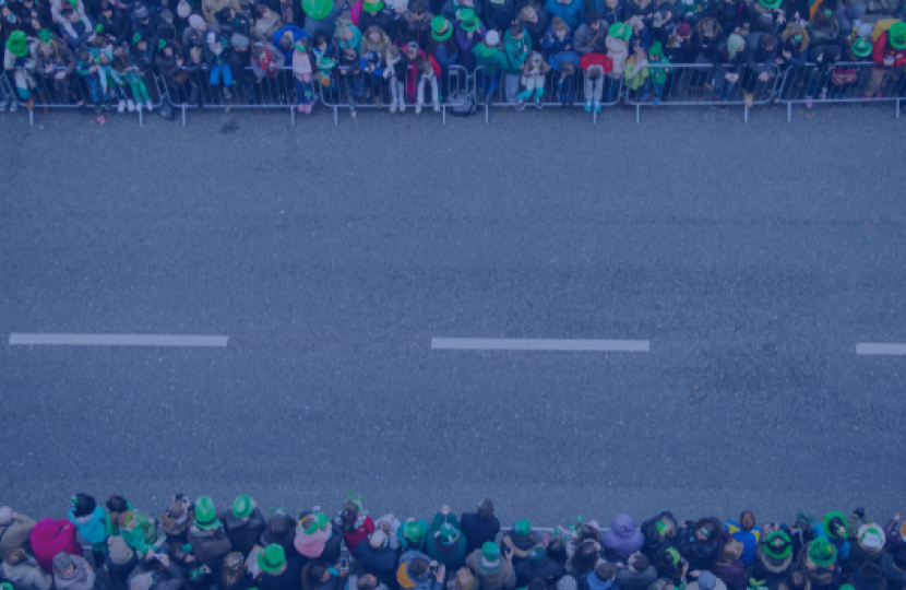 A road prepared for a parade. People gathered either side behind barriers wearing Irish themed paraphernalia 