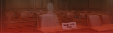 A ghostly figure sits in an empty council chamber